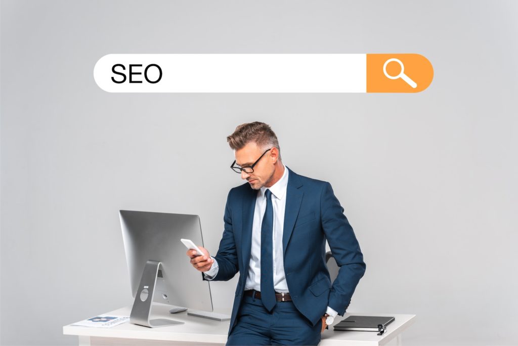 handsome businessman leaning on table and using smartphone isolated on grey with seo in search bar