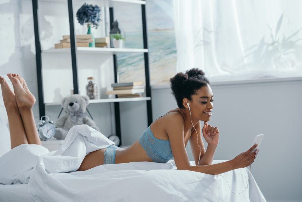 happy african american girl in lingerie listening music with earphones and smartphone in bedroom in