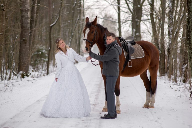 Happy newly married couple standing with beautiful brown horse in winter nature